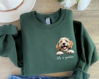 以下が含まれることがあります： 笑顔のゴールデン・ドゥードゥル犬のグラフィックと「life is golden」の文字が入った濃い緑色のスウェットシャツ。袖は折り返されています。茶色のバンドが付いた白い帽子が背景に見えます。スウェットシャツは折りたたまれています。