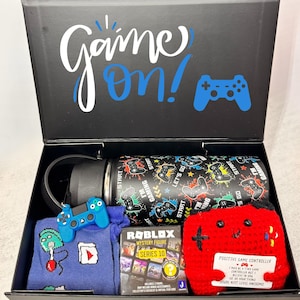 May include: A black gift box with the words "Game On!" and a blue controller graphic. Inside are a black and multi-colored water bottle, a blue controller keychain, a blue sweater, a Roblox mystery figure box, and a red crocheted controller.