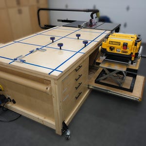 Puede incluir: Un banco de trabajo de madera con una cepilladora DEWALT amarilla, una sierra de mesa y una manguera de aspiración de polvo azul. El banco de trabajo tiene cuatro cajones y está sobre ruedas. La parte superior del banco de trabajo tiene cinta azul marcando un patrón de cuadrícula.