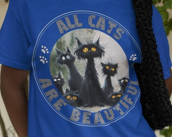 May include: A woman wearing a blue t-shirt with a white circle graphic featuring the text "All Cats Are Beautiful" and a cartoon image of four black cats.