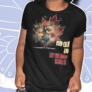 Könnte beinhalten: Schwarzes T-Shirt mit einer Grafik eines Cartoon-Charakters, der ein Ahornblatt, einen roten Hut und eine Aktentasche hält. Der Text "Keep Calm and Let the Moose Handle It" ist auf dem Shirt aufgedruckt. Der Hintergrund ist hellblau mit einem Schmetterlingsdesign.