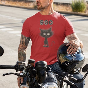 May include: A man wearing a red t-shirt with a black cat graphic and the word "BOO" printed on it. He is sitting on a motorbike.