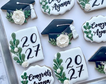 Può includere: Biscotti a tema laurea disposti su un vassoio. I biscotti presentano cappelli neri da laurea, l'anno 2027 e la scritta "Congrats Ayana". Ogni biscotto è decorato con fiori bianchi e foglie verdi.