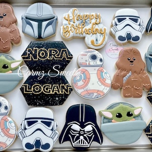 May include: A set of Star Wars themed sugar cookies decorated with various characters, including Yoda, Darth Vader, Stormtroopers, and BB-8. The cookies are decorated with black, white, brown, and orange icing. The cookies are arranged on a white surface. The cookies are labeled with the names "Nora" and "Logan".