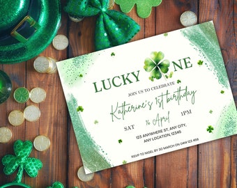 Può includere: Un invito verde e bianco con un design a trifoglio e il testo "LUCKY ONE" e "Katherine's 1st birthday". L'invito è su una superficie di legno con decorazioni a trifoglio verde.