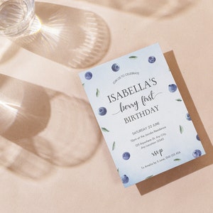 May include: A blue and white invitation card with blueberries and leaves printed on it. The text on the card reads "Join us to celebrate Isabella's berry first birthday. Saturday 22 June 12pm at the Jacobs Residence 123 Anywhere St, Any City, Any Location 12345. rsvp to Amelia by 5 June 0141 123 456"