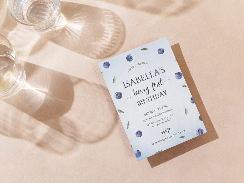 May include: A blue and white invitation card with blueberries and leaves printed on it. The text on the card reads "Join us to celebrate Isabella's berry first birthday. Saturday 22 June 12pm at the Jacobs Residence 123 Anywhere St, Any City, Any Location 12345. rsvp to Amelia by 5 June 0141 123 456"