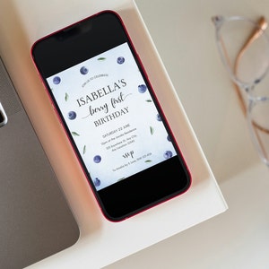 May include: A smartphone displaying a digital invitation for Isabella's first birthday party. The invitation features a blueberry theme with the text "Isabella's berry first birthday" and a date and time for the event. The invitation is on a white background with a pattern of blueberries and green leaves.