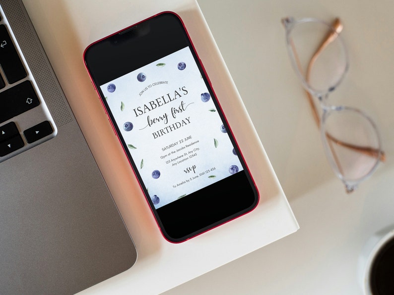 May include: A smartphone displaying a digital invitation for Isabella's first birthday party. The invitation features a blueberry theme with the text "Isabella's berry first birthday" and a date and time for the event. The invitation is on a white background with a pattern of blueberries and green leaves.
