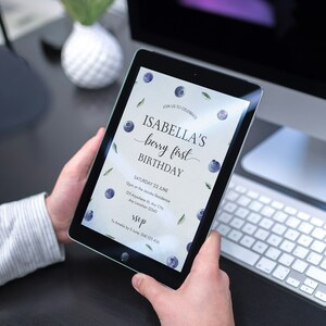 May include: A digital tablet displaying a blue and green birthday invitation with the text "JOIN US TO CELEBRATE ISABELLA'S berry first BIRTHDAY SATURDAY 22 JUNE 123 Anywhere St Any City Any Location 12343 RSVP 14 Amelia Dr, 5 June, 0123 456"