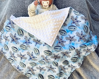 Op de afbeelding: Een baby draagt ​​een witte muts met het woord "COWBOYS" erop en ligt op een blauw-witte deken met een witte minky-rug. De deken heeft een patroon van sterren en voetballen met het woord "COWBOYS" dat overal wordt herhaald.