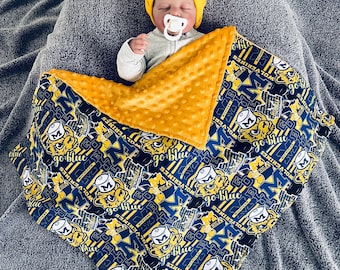 May include: A baby wearing a yellow beanie and a pacifier, wrapped in a blue and yellow blanket. The blanket features a repeating pattern with the text "Wolverines" and "Go Blue" along with a cartoon tiger mascot.