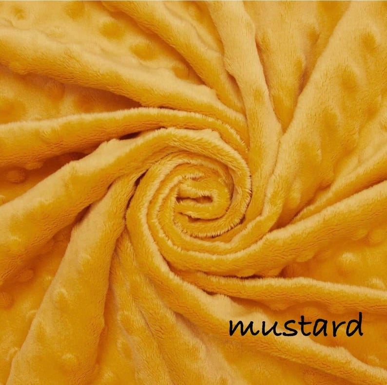May include: Mustard-colored fabric with a soft, textured surface. The fabric is swirled, creating a spiral pattern. The word "mustard" is printed in black at the bottom right corner. The fabric has a plush, dotted texture.