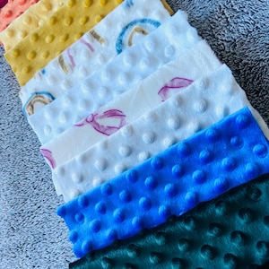 May include: A collection of colorful, textured fabric squares. The squares are in shades of burgundy, peach, yellow, white, blue, and green. Some squares have a rainbow or bow design. The fabric has a bumpy, dotted texture.