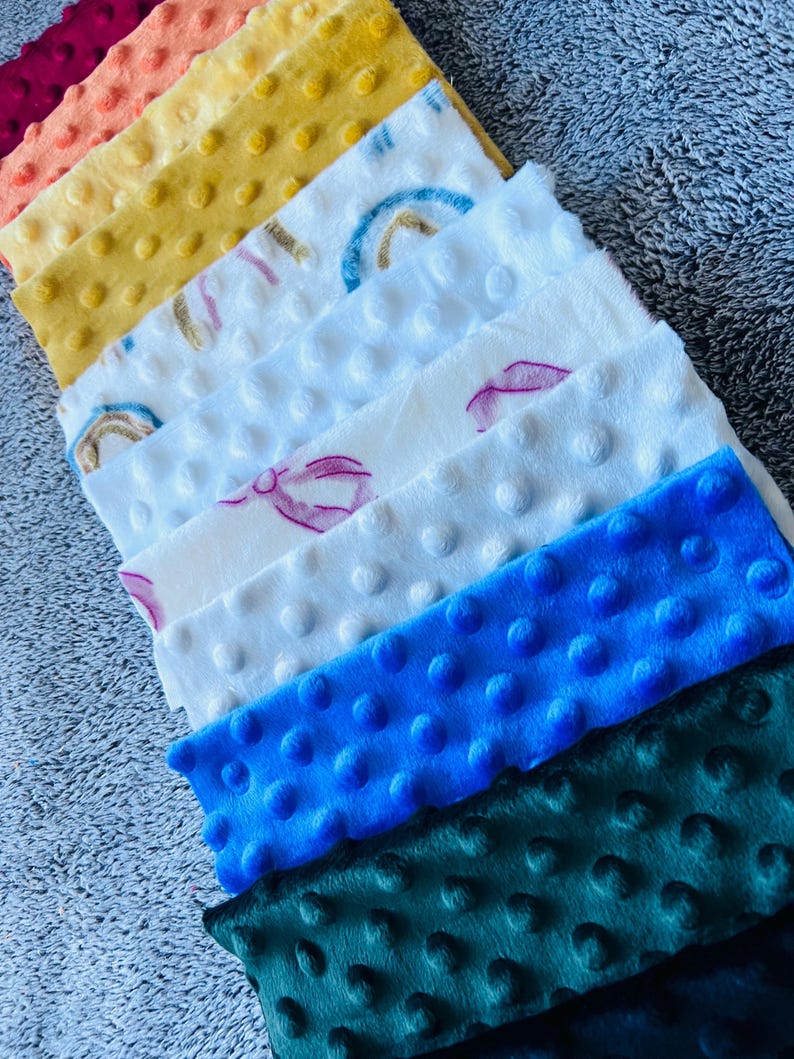 May include: A collection of colorful, textured fabric squares. The squares are in shades of burgundy, peach, yellow, white, blue, and green. Some squares have a rainbow or bow design. The fabric has a bumpy, dotted texture.