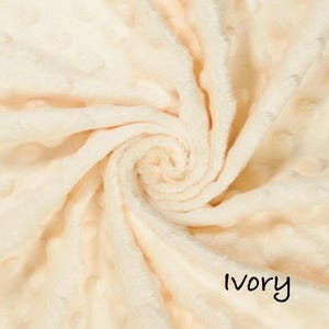 May include: Ivory-colored, plush fabric with a swirl pattern. The soft material has a textured surface with small, raised dots. The word "Ivory" is written in a black, handwritten font at the bottom right of the image. This fabric is suitable for blankets and other soft goods.