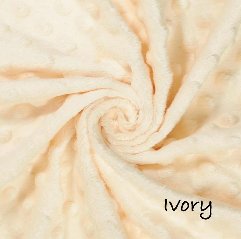 May include: Ivory-colored, plush fabric with a swirl pattern. The soft material has a textured surface with small, raised dots. The word "Ivory" is written in a black, handwritten font at the bottom right of the image. This fabric is suitable for blankets and other soft goods.