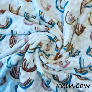 May include: White, textured blanket with a repeating rainbow pattern in shades of brown, pink, and blue. The word "rainbow" is printed in black script at the bottom right corner. The blanket is soft and plush.