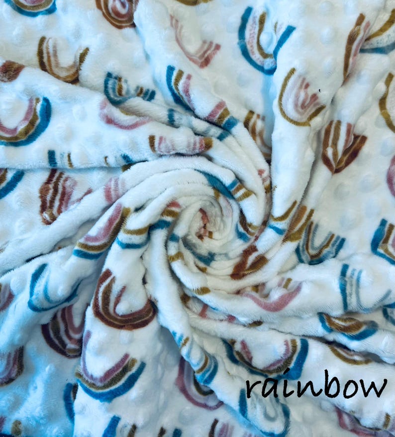 May include: White, textured blanket with a repeating rainbow pattern in shades of brown, pink, and blue. The word "rainbow" is printed in black script at the bottom right corner. The blanket is soft and plush.