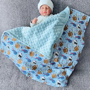 May include: A baby wrapped in a light blue blanket with a football-themed pattern. The blanket features brown footballs, helmets, and goalposts on a light blue background. The top layer is a soft, textured, light blue blanket.