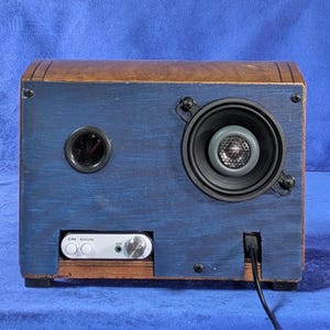 May include: A handmade wooden speaker with a blue painted front and a brown top. The speaker has a round speaker, a small round port, and a white audio input panel. A black power cord extends from the bottom.