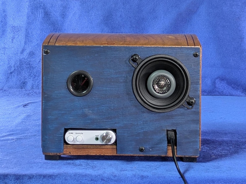 May include: A handmade wooden speaker with a blue painted front and a brown top. The speaker has a round speaker, a small round port, and a white audio input panel. A black power cord extends from the bottom.