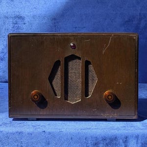 May include: A vintage wooden radio with a dark brown finish. The radio features a speaker grill, two knobs, and a small purple indicator light. The radio is rectangular and sits on a blue surface.