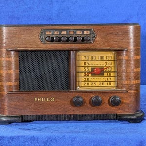 May include: A vintage Philco radio with a dark wood finish. The radio features a speaker grill, a tuning dial, and several knobs. The dial displays frequency numbers. The word "PHILCO" is printed on the front.