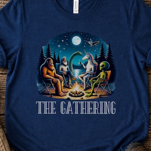 K&ouml;nnte beinhalten: Ein marineblaues T-Shirt mit einem Grafikdruck von Bigfoot, einem b&auml;rtigen Mann, einem Einhorn und einem Alien, die nachts unter einem Vollmond um ein Lagerfeuer sitzen. Der Text THE GATHERING ist in wei&szlig;en Gro&szlig;buchstaben darunter gedruckt.