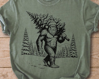 Könnte beinhalten: Ein salbeigrünes Kurzarm-T-Shirt mit einem schwarzen Grafikdruck eines Bigfoot-Wesens, das eine Weihnachtsmütze trägt und einen Tannenbaum über der Schulter trägt. Das Design zeigt einen Waldhintergrund mit mehreren Tannenbäumen und Bergen.