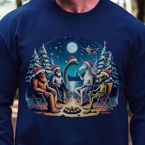 May include: A navy blue long-sleeve sweatshirt featuring a graphic print of Bigfoot, a bearded person, the Loch Ness Monster, a unicorn, and an alien sitting around a campfire. Each character wears a red Santa hat in a snowy forest setting.