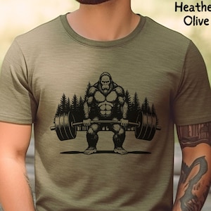 Heather olive green t-shirt featuring a black graphic of a Bigfoot lifting a barbell with trees in the background. The shirt is made of a soft material.
