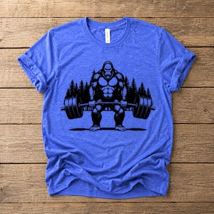 Heather true royal t-shirt featuring a black graphic of a Bigfoot lifting a barbell. The design includes a forest backdrop.