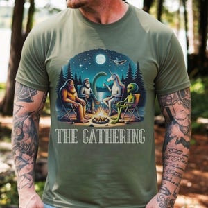K&ouml;nnte beinhalten: Ein olivgr&uuml;nes T-Shirt mit einem Grafikdruck von Bigfoot, einer b&auml;rtigen Person, einem Einhorn und einem Alien, die nachts um ein Lagerfeuer sitzen. Der Text "THE GATHERING" ist in wei&szlig;en Gro&szlig;buchstaben unter der farbigen Illustration gedruckt.