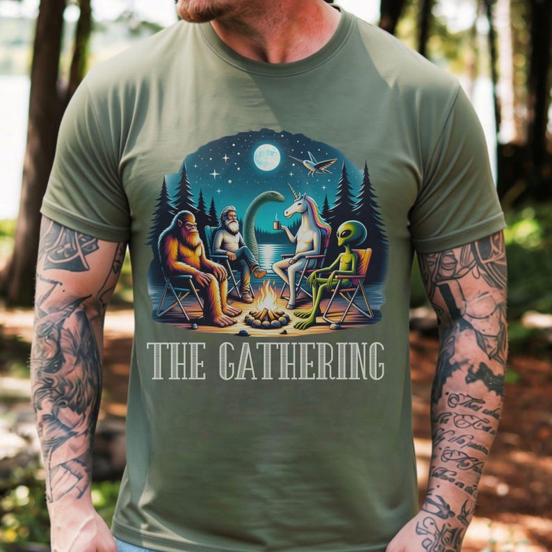 K&ouml;nnte beinhalten: Ein olivgr&uuml;nes T-Shirt mit einem Grafikdruck von Bigfoot, einer b&auml;rtigen Person, einem Einhorn und einem Alien, die nachts um ein Lagerfeuer sitzen. Der Text "THE GATHERING" ist in wei&szlig;en Gro&szlig;buchstaben unter der farbigen Illustration gedruckt.