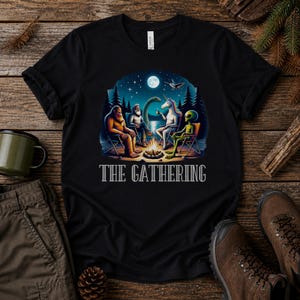 K&ouml;nnte beinhalten: Ein schwarzes T-Shirt mit einer Grafik von Bigfoot, einer b&auml;rtigen Person, einem Einhorn und einem Alien, die nachts um ein Lagerfeuer sitzen. Der Text THE GATHERING ist in Wei&szlig; unter der Illustration gedruckt, die einen See und Vollmond zeigt.