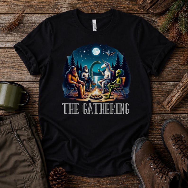 K&ouml;nnte beinhalten: Ein schwarzes T-Shirt mit einer Grafik von Bigfoot, einer b&auml;rtigen Person, einem Einhorn und einem Alien, die nachts um ein Lagerfeuer sitzen. Der Text THE GATHERING ist in Wei&szlig; unter der Illustration gedruckt, die einen See und Vollmond zeigt.