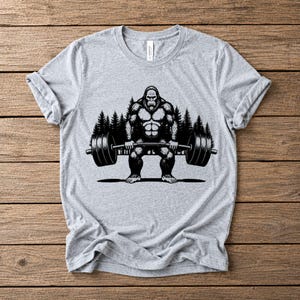 Athletic heather t-shirt featuring a black graphic of a Bigfoot lifting a barbell. The design includes a forest backdrop.