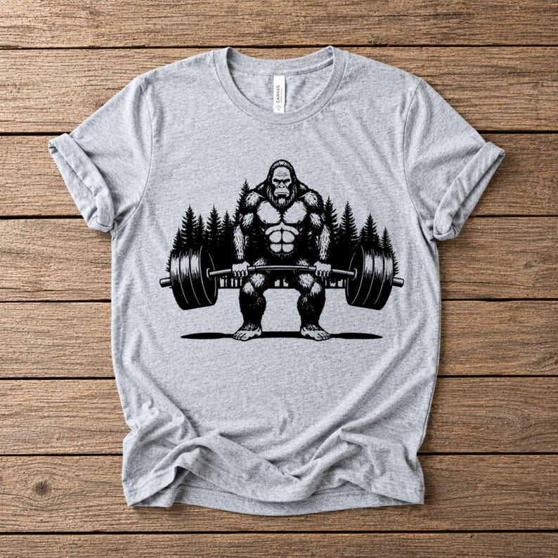Athletic heather t-shirt featuring a black graphic of a Bigfoot lifting a barbell. The design includes a forest backdrop.