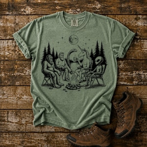 Moss Comfort Colors t-shirt featuring a black ink art design of The Gathering of Friends. Bigfoot, unicorn, alien, Loch Ness Monster and the mysterious man spending time together around a campfire. Set in wooded scene on a lake at nighttime.