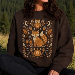 May include: A dark brown crewneck sweatshirt featuring a central graphic print of a fox surrounded by an owl, a pumpkin, acorns, birds, crescent moons, stars, and oak leaves in shades of orange, cream, and black, with long sleeves and ribbed trim.