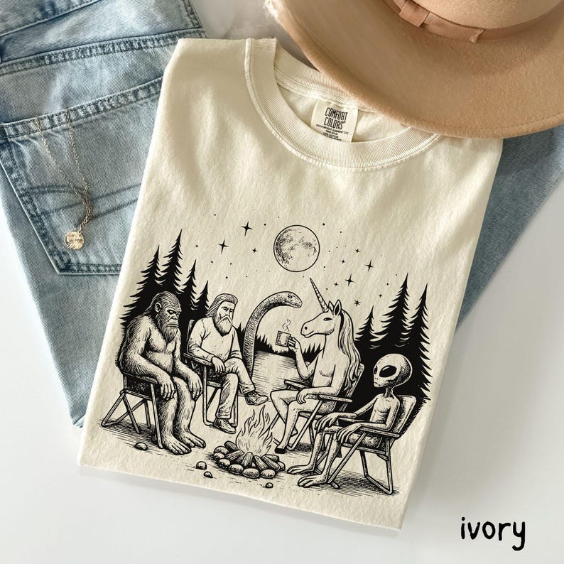 Ivory Comfort Colors t-shirt featuring a black ink art design of The Gathering of Friends. Bigfoot, unicorn, alien, Loch Ness Monster and the mysterious man spending time together around a campfire. Set in wooded scene on a lake at nighttime.