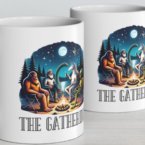 May include: Two white ceramic mugs with a colorful illustration of a group of mythical creatures gathered around a campfire. The text "The Gathering" is printed on both mugs.