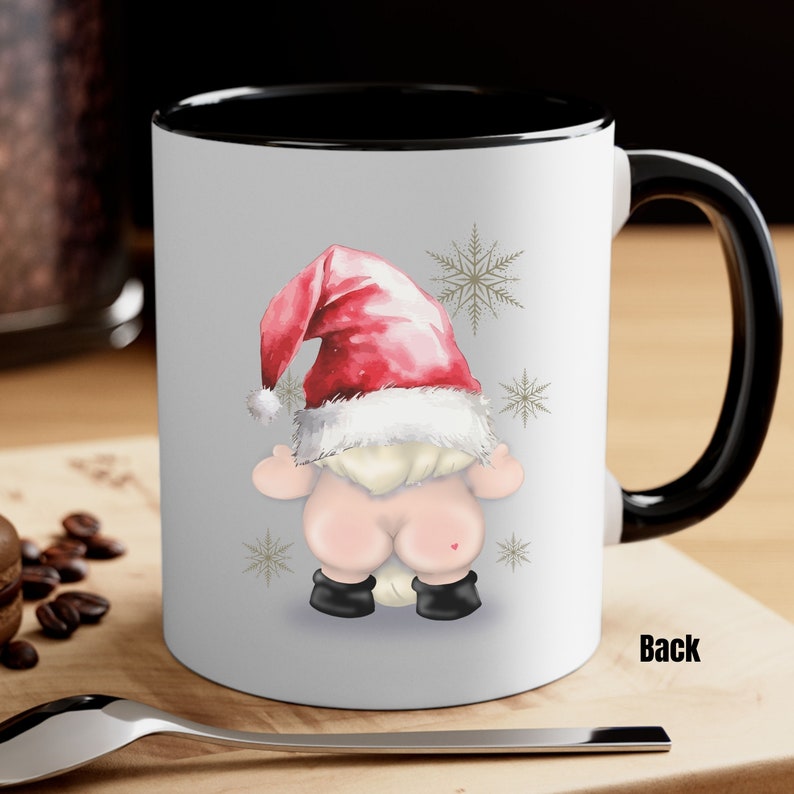 May include: White ceramic mug with a black rim and handle. The mug features a cartoon illustration of a gnome's backside wearing a red and white Santa hat. The gnome is surrounded by snowflakes.
