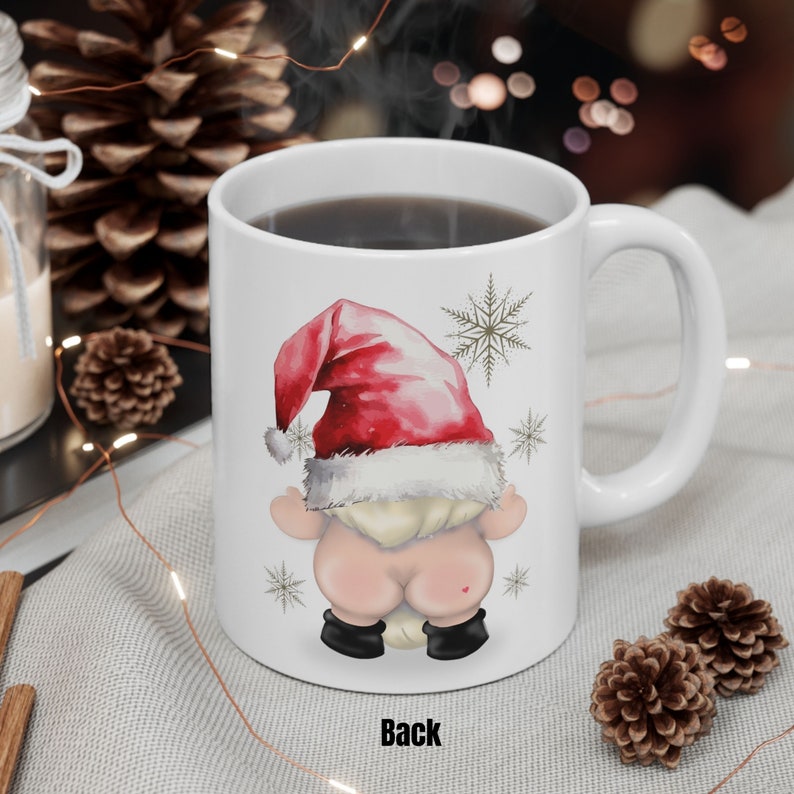 May include: White ceramic coffee mug featuring a red and white Santa hat gnome design. The gnome is facing away from the viewer with its back to the camera. The mug has the word "Back" printed on the bottom.