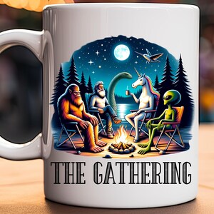 May include: White ceramic mug with a colorful illustration of a Bigfoot, a unicorn, an alien, and a man with a beard sitting around a campfire under a starry night sky. The text "THE GATHERING" is printed in black at the bottom of the mug.