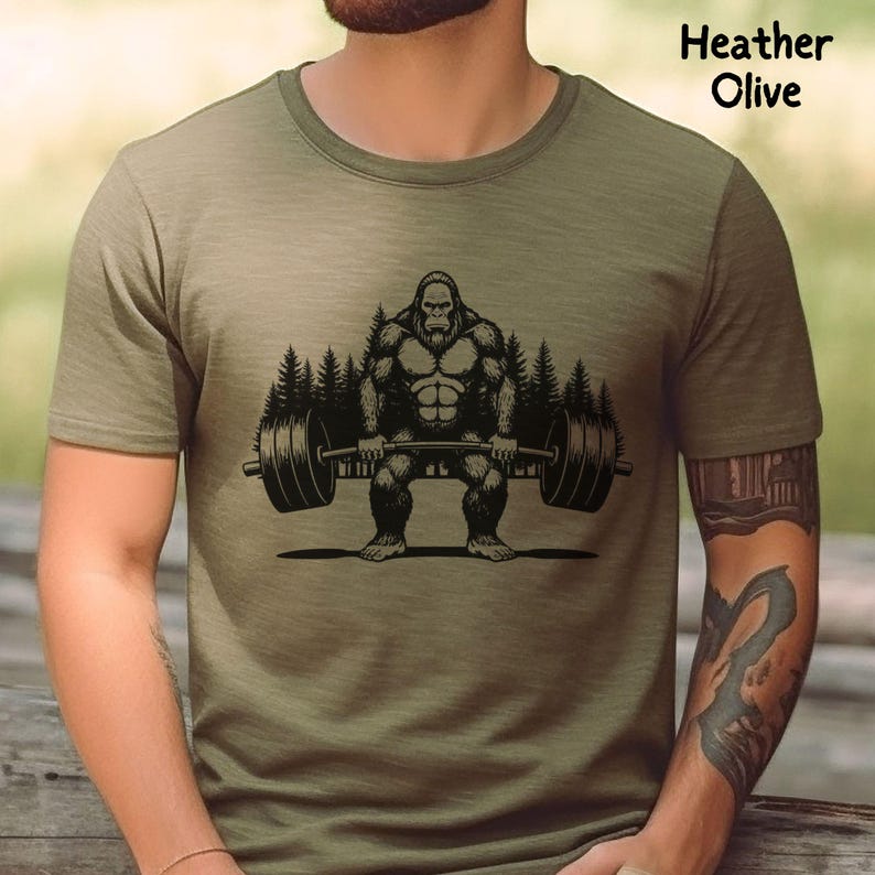 Heather olive green t-shirt featuring a black graphic of a Bigfoot lifting a barbell with trees in the background. The shirt is made of a soft material.