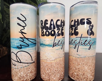 Op de afbeelding: Drie roestvrijstalen bekers met een strandscène en tekst. De eerste beker zegt "Brynn", de tweede zegt "Beach Booze & Besties" en de derde zegt "Beaches & Besties".
