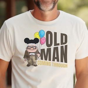 May include: White t-shirt with a cartoon illustration of an older man with glasses wearing a Mickey Mouse hat and holding balloons. The text "OLD MAN COMING THROUGH" is printed on the shirt.