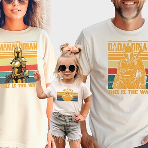 May include: A set of three white t-shirts with a retro-style graphic of a Mandalorian character. The shirts have the text "The Mamadorean", "The Dadadorean", and "The Child" with the phrase "This is the way" printed below the graphic.